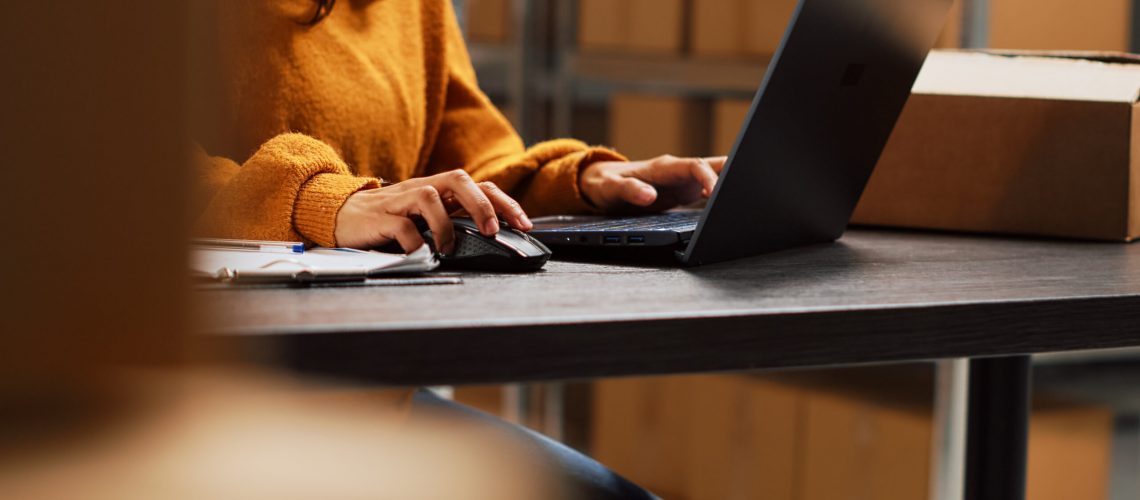 Startup owner working on stock inventory with laptop, planning order distribution for business management in storage room. Worker doing supply chain shipment at desk, retail products. Handheld shot.
